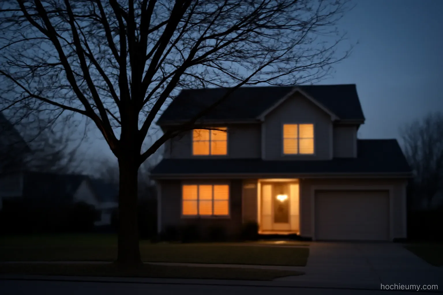 a house with lights on seen from the street at dusk with a bare tree standing in the front yard